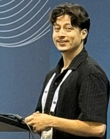 A person standing near a podium, smiling, set against a blue background with white wave patterns.