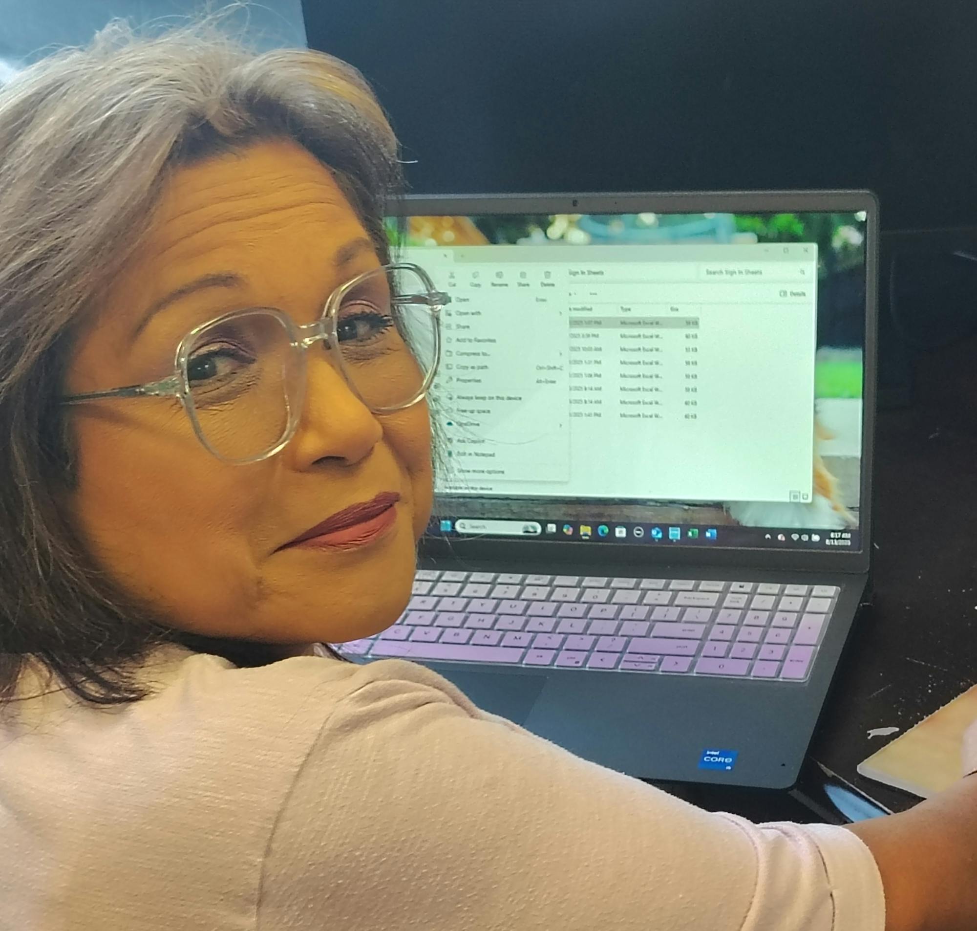 A person with glasses is seated at a desk, looking at a laptop displaying a spreadsheet or data.