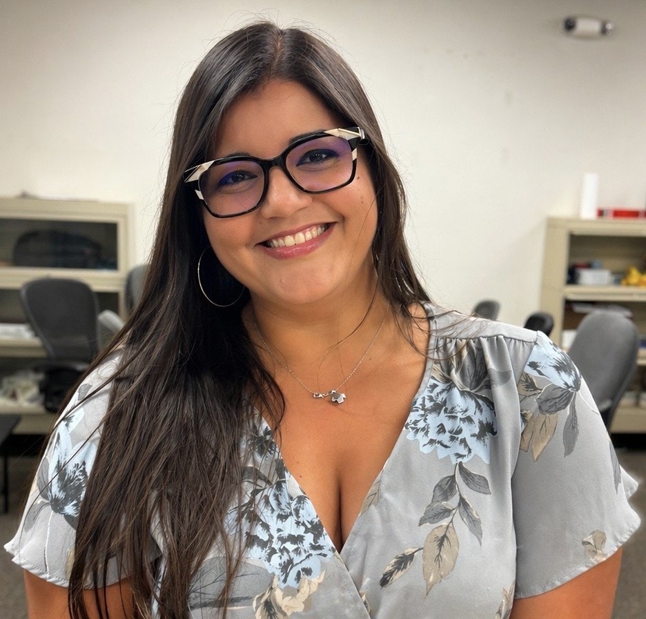 A woman with long hair and glasses smiles warmly, wearing a floral top in a bright office setting.