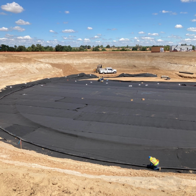 Large excavation area with geotextile liner being installed, construction equipment, and workers visible, under a clear sky.