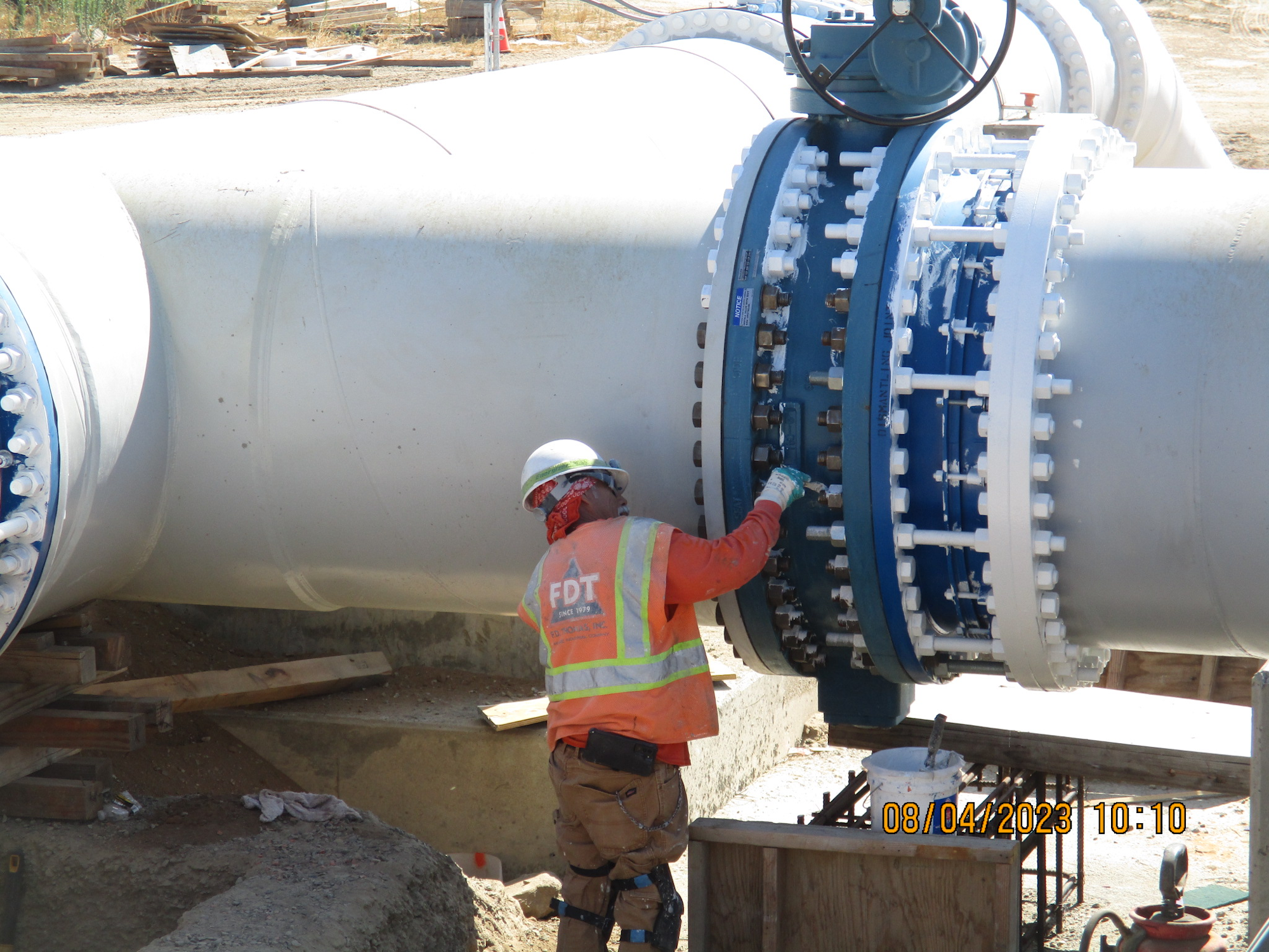 A worker with a hardhat inspects a large pipeline with flanged joints at a construction site.