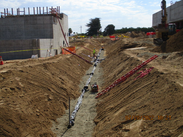 A construction site with a trench, pipework, and concrete structures, under a cloudy sky.