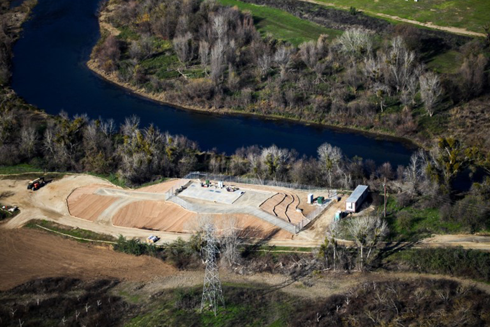 An aerial view of a construction site near a river, with machinery, structures, and terraced layers of earth.
