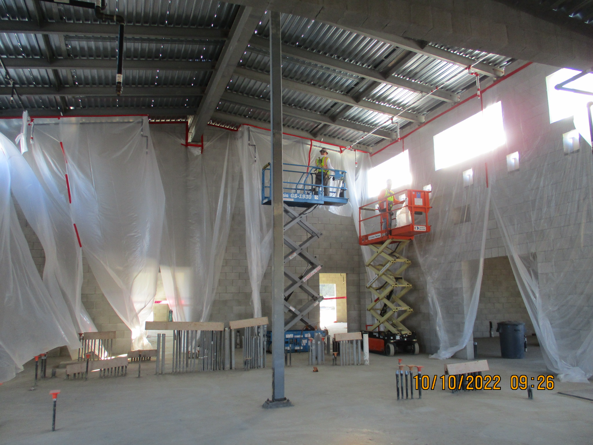 Industrial interior with workers on scissor lifts, plastic sheeting, metal beams, and construction materials.