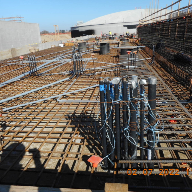 Construction site with steel rebar framework, conduit pipes, and workers in safety gear.