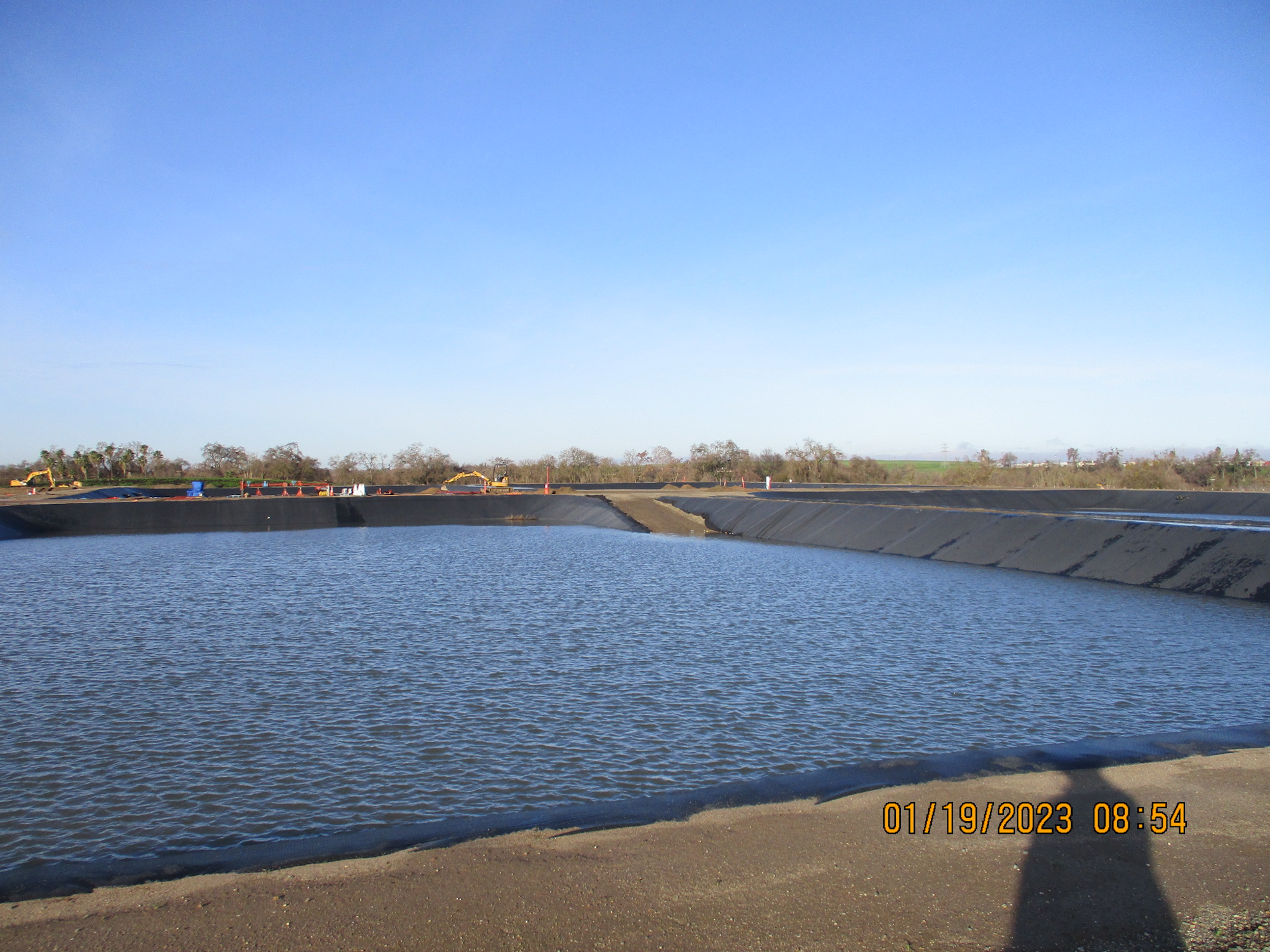 A large water reservoir with reinforced edges under a clear blue sky, construction equipment and materials in the background.
