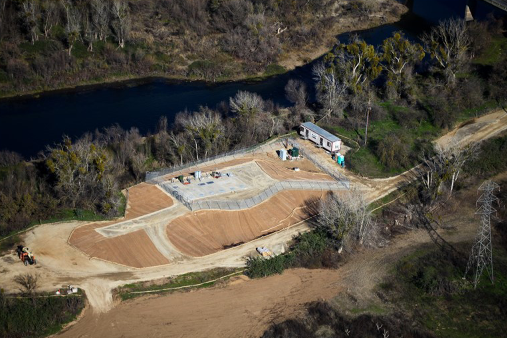 An aerial view of a construction site near a river with groundwork laid out, machinery, and a few buildings.