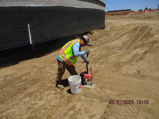 Construction worker using a compacting machine on soil near a large tank, with safety gear on, under clear skies.