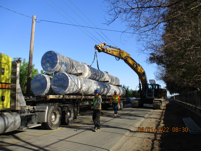 An excavator lifting large coils onto a truck, with workers supervising.