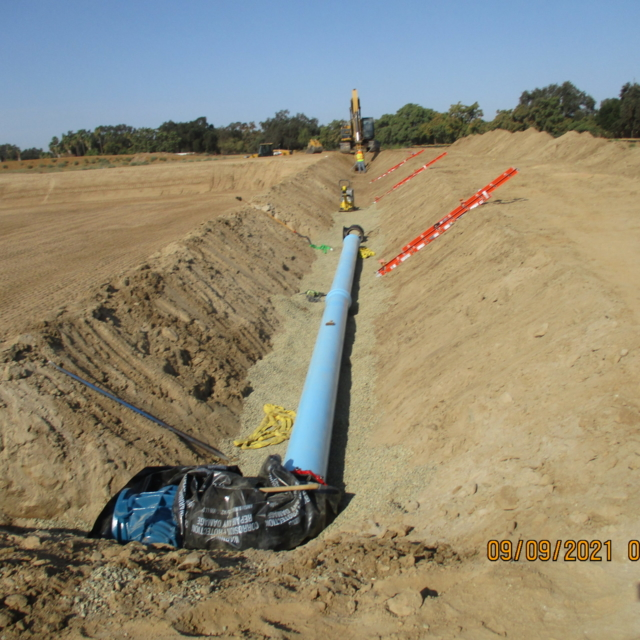 A blue pipeline being installed in a trench with construction equipment and workers in the background.
