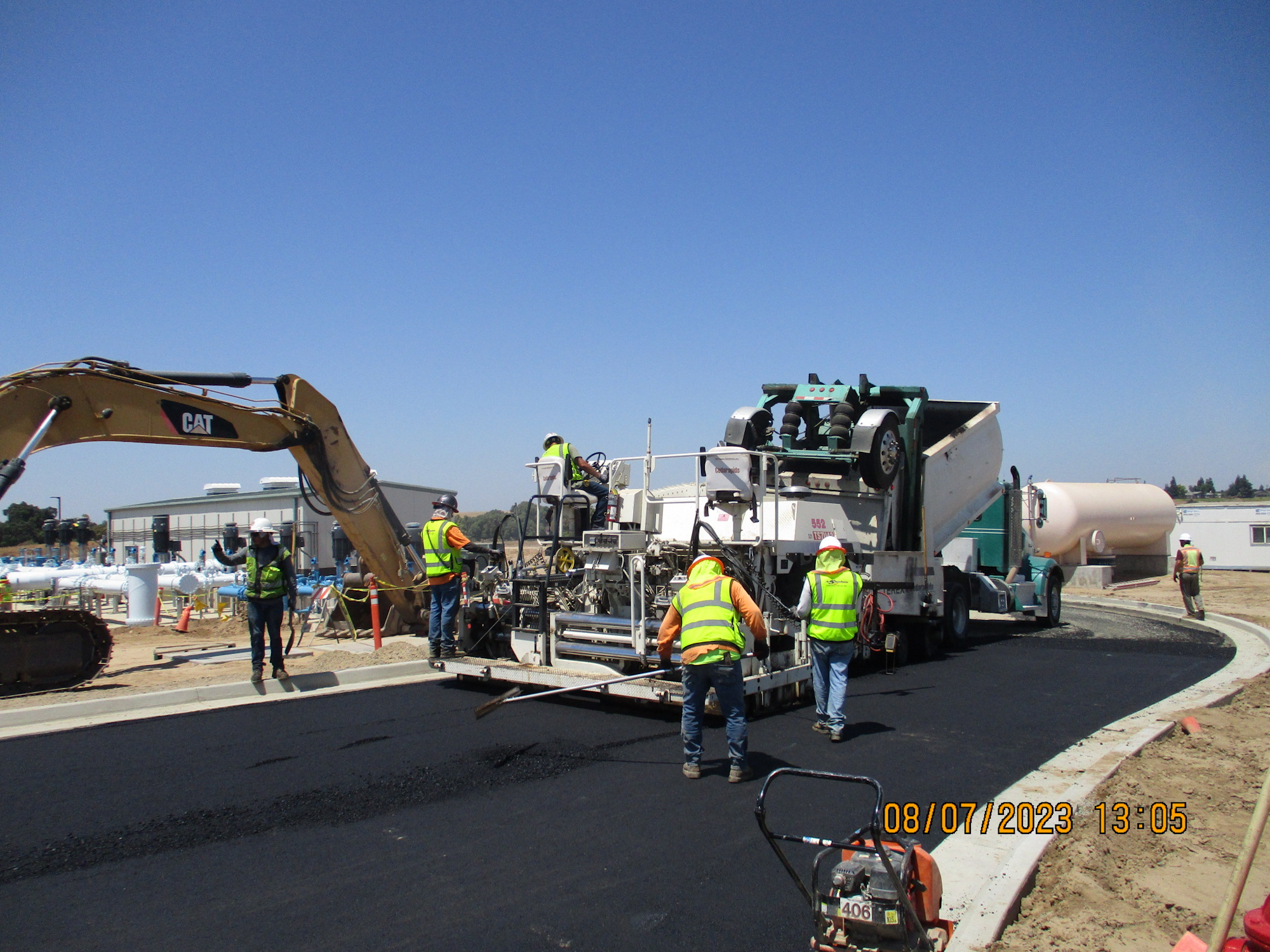 Workers in high-visibility vests operate machinery for road construction under a clear sky.
