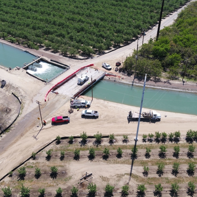 Aerial view of a waterway, with vehicles by a road, trees in rows, and construction or maintenance activity near a gate structure.