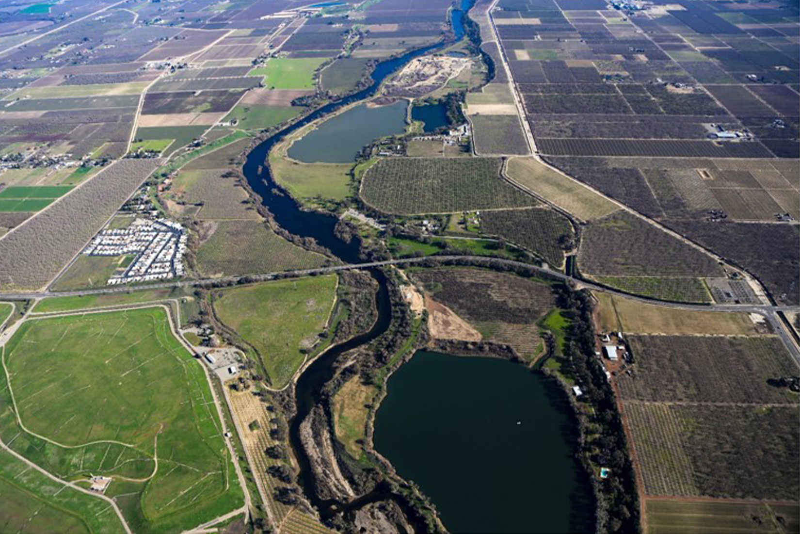 Aerial view of a winding river flanked by agricultural fields, with roads and a circular pond.