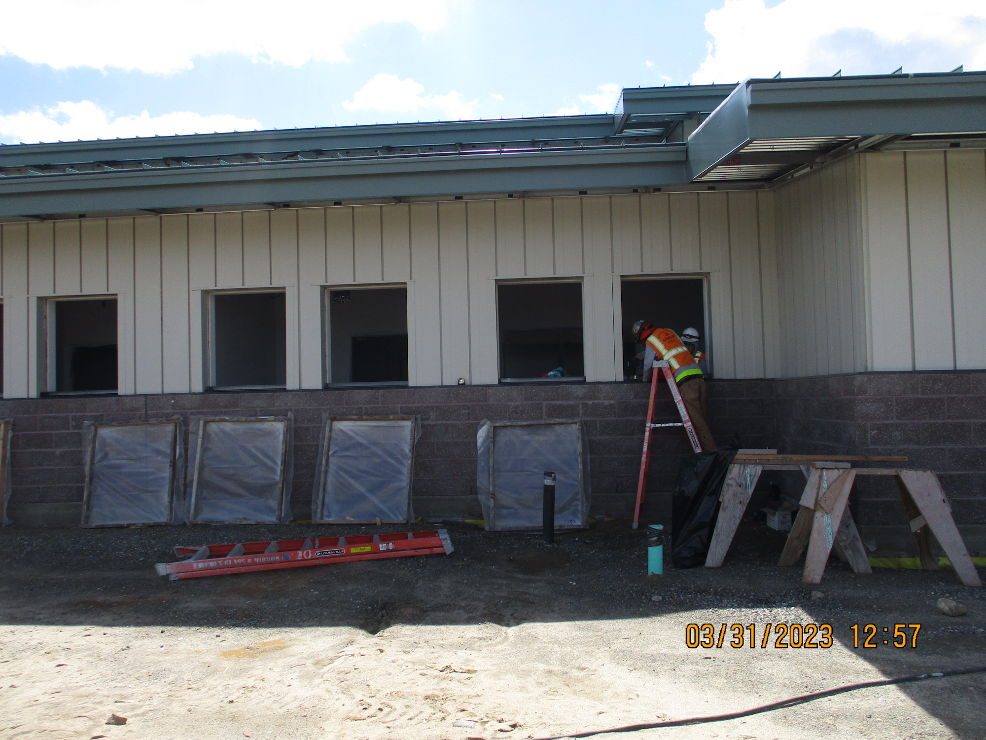 Construction workers on a ladder by a building with open windows, tools, and materials on the ground, under a clear sky.