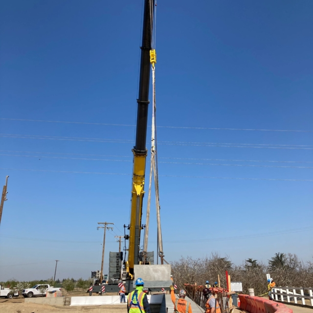 A crane lifting a large concrete panel with construction workers guiding it on a construction site, under a clear blue sky.
