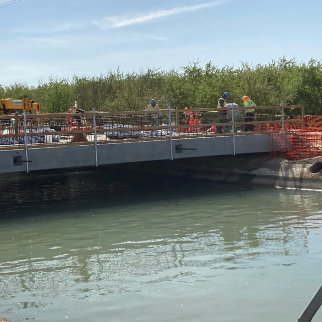 Construction workers on a bridge over water with vegetation in the background.