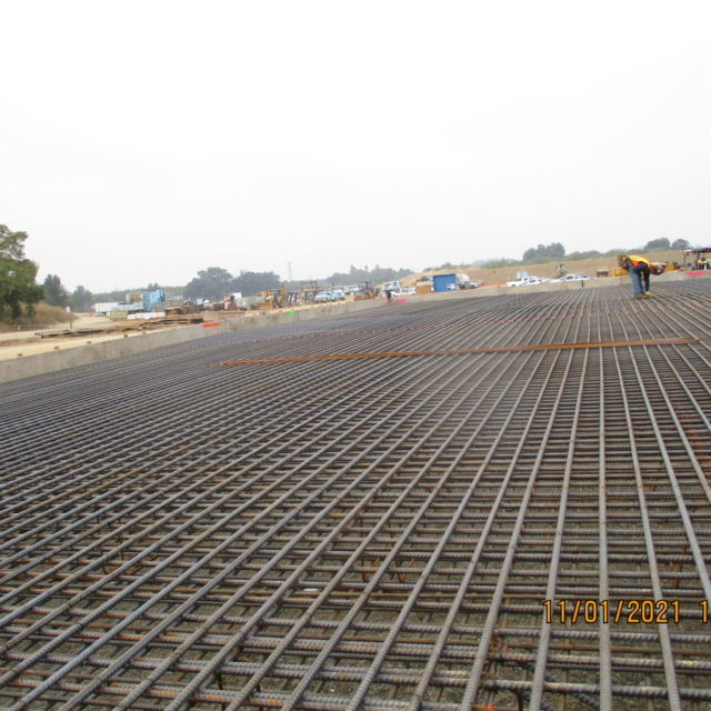 Construction site with steel rebar grid, workers, and equipment in background.