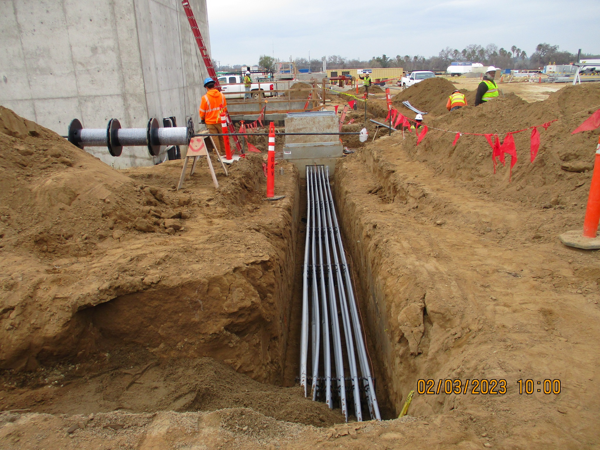 A construction site with workers, an open trench with pipes, and safety barriers.