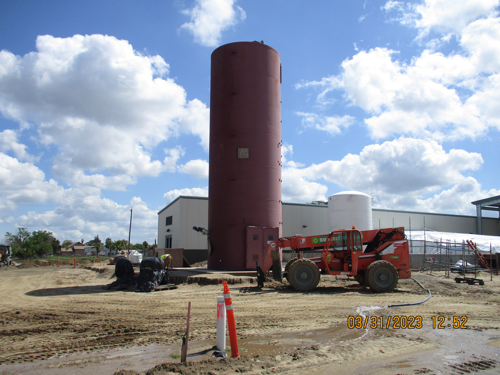Construction site with a telescopic handler, silo, and building under blue sky with clouds.