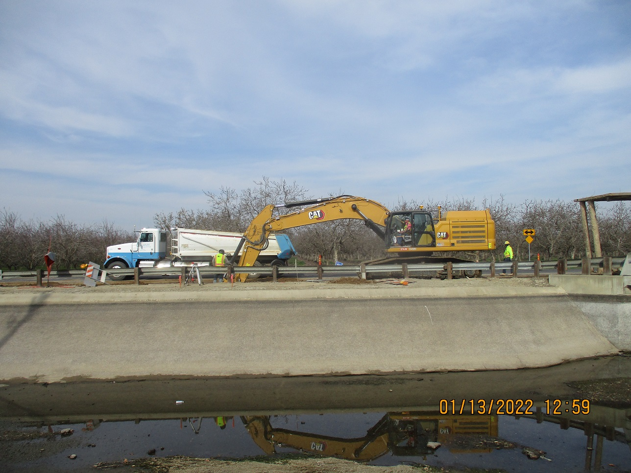 Construction site with an excavator, truck, workers, concrete barriers, and trees, with a water reflection at the bottom.