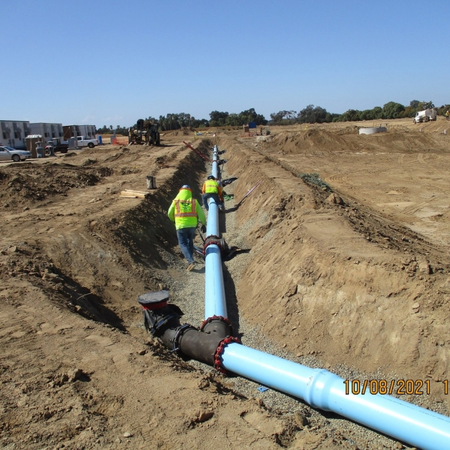 Construction workers inspect a large pipeline in a trench with construction and development in the background.