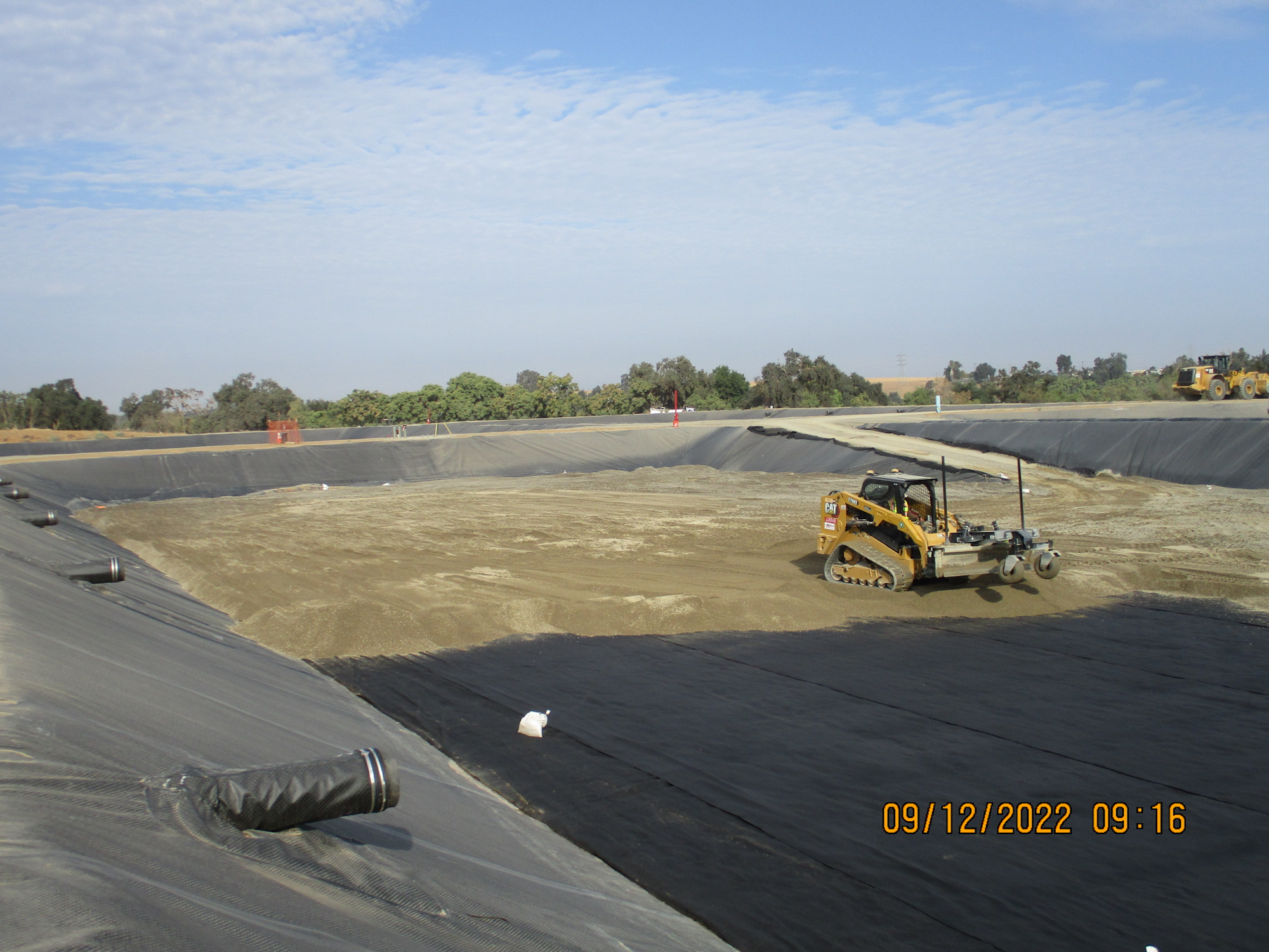 Construction site with heavy machinery, lined with black geotextile fabric, under a clear sky.