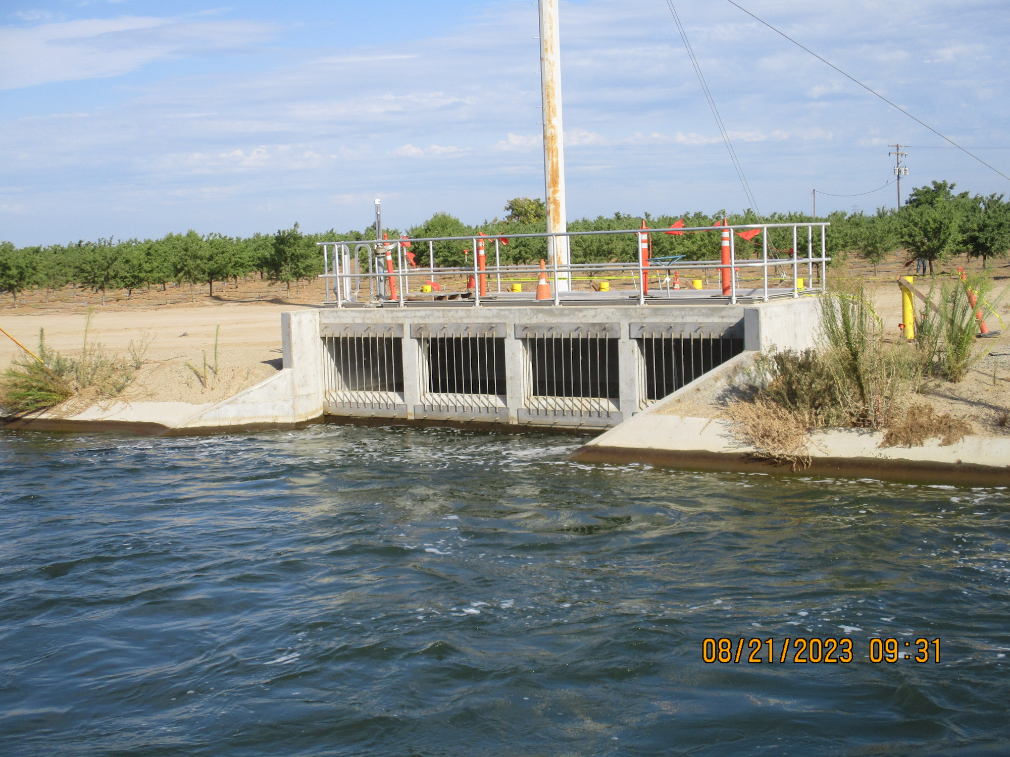 Water control structure with gates, surrounded by fencing, near agriculture area, with timestamp.