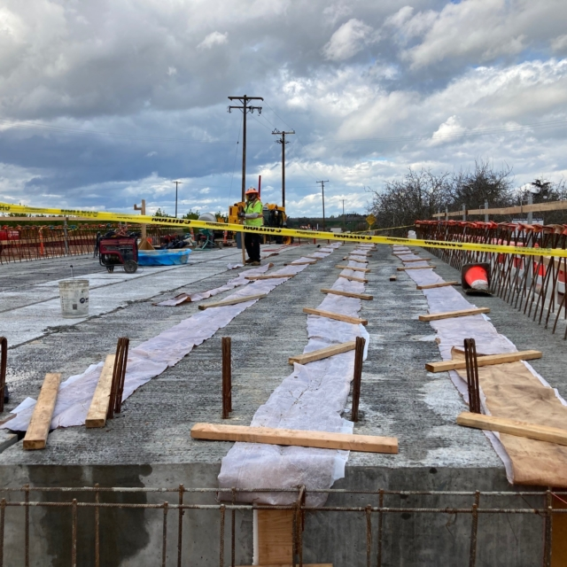 Construction site with concrete foundation, wooden planks, fabric sheets, and a worker surrounded by caution tape.