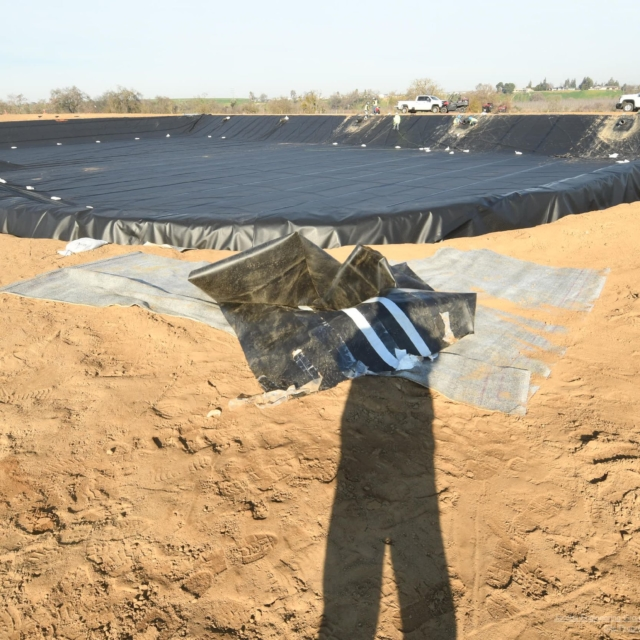 A damaged plastic liner on dirt with a person's shadow in the foreground.
