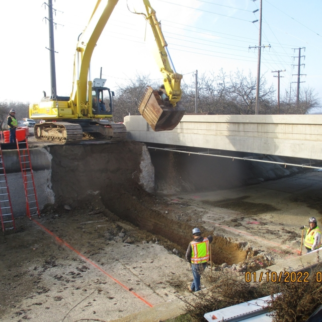 Excavator digging near a bridge with workers overseeing.