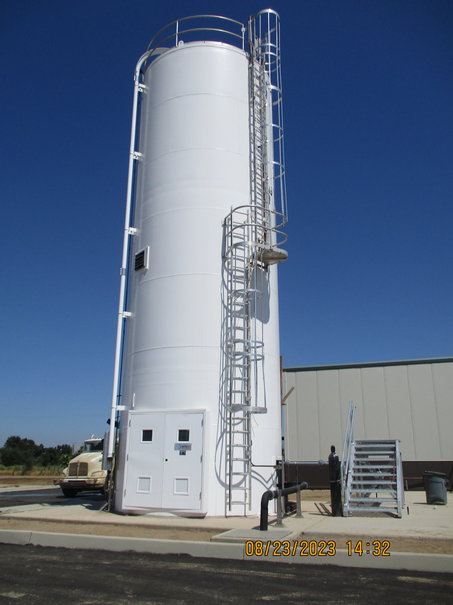 A large white storage tank with a staircase, under a clear sky, with a person and a truck nearby.
