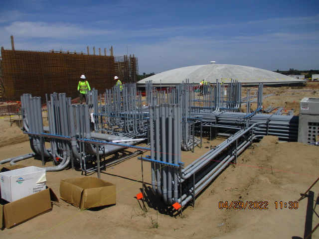 Construction site with conduit pipes, workers in the background, and a partially built structure under a blue sky.
