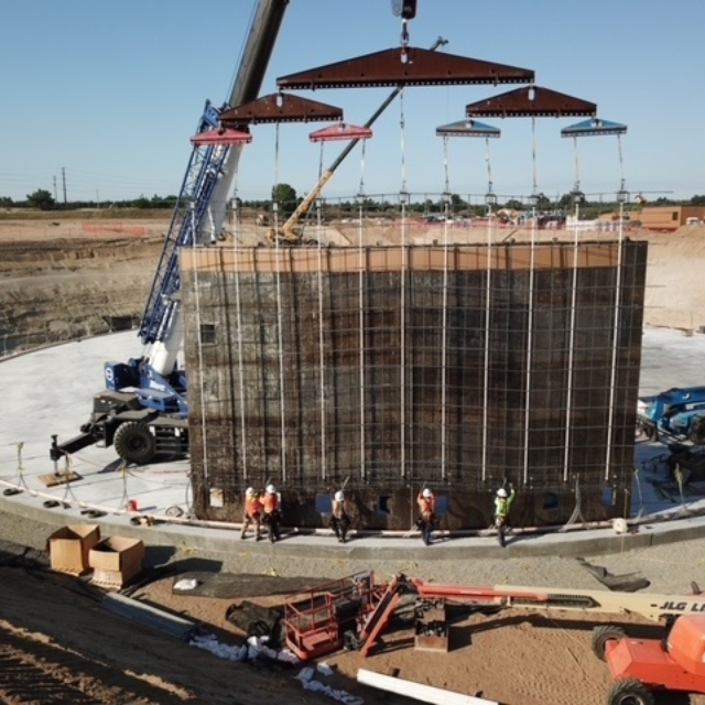 Construction site with workers, a crane lifting materials, and a large cylindrical concrete formwork.