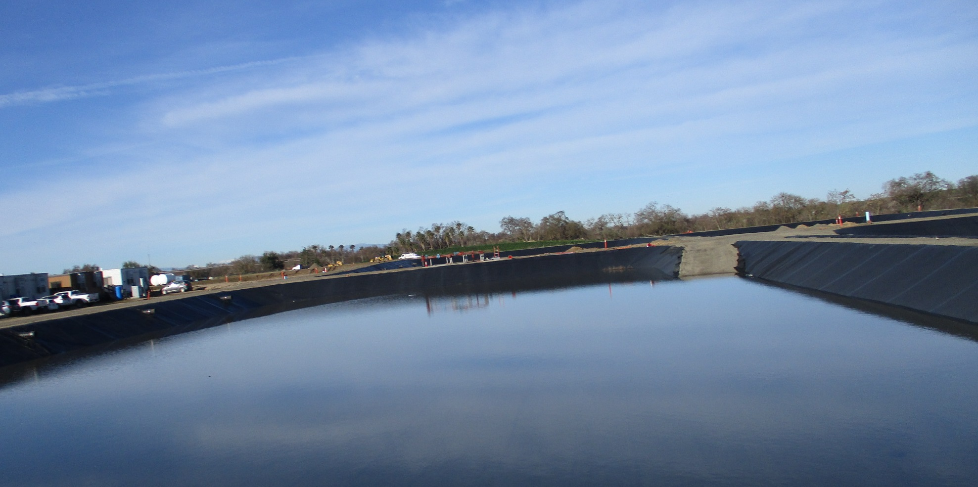 Large water retention pond with black liner and industrial equipment in the background under a clear blue sky.