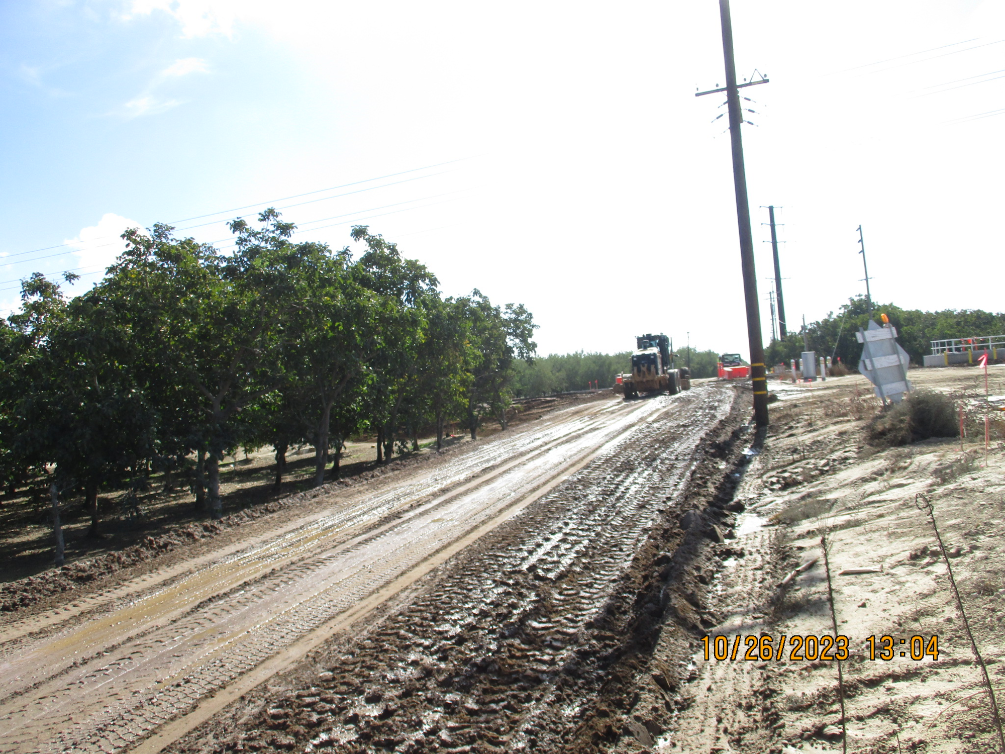 A muddy construction site with machinery, utility poles, and trees under a clear sky.