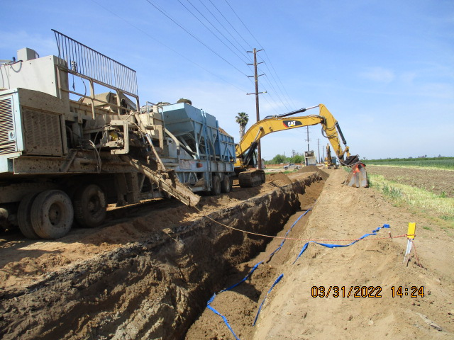 Construction equipment digging a trench with workers on a sunny day.