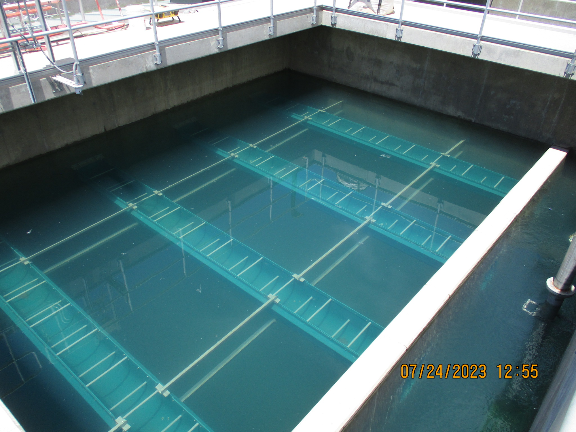 A water treatment or storage basin with metal bars across it, date stamped with time "07/24/2023 12:55".
