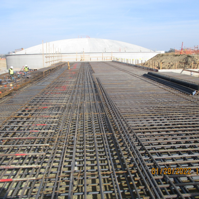 A construction site with reinforcement bar framework laid out for concrete, with a large dome structure in the background.