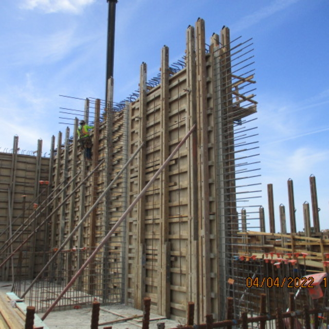 A construction site with wooden formwork and steel reinforcement bars. A worker is visible on the structure.
