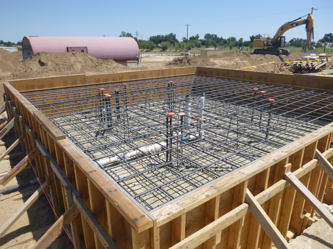 Construction site with foundation framework, rebar grid, and plumbing, before concrete pour. Excavator in background.