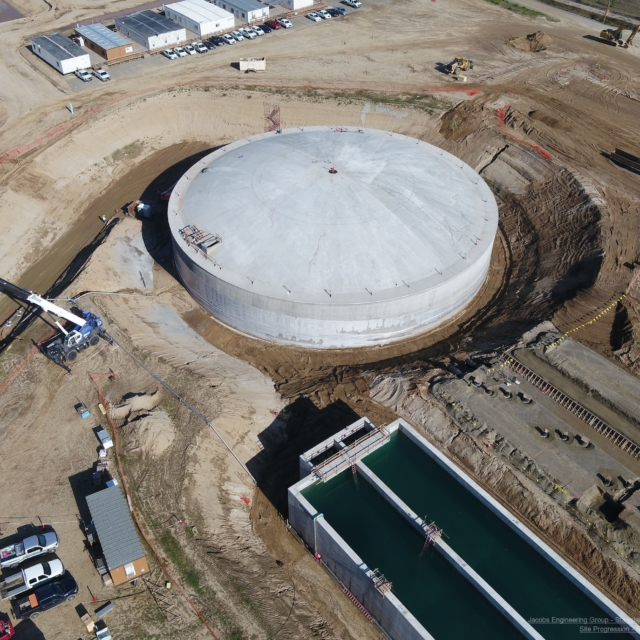 Aerial view of a large circular tank under construction with an adjacent rectangular basin, surrounded by construction vehicles and equipment.
