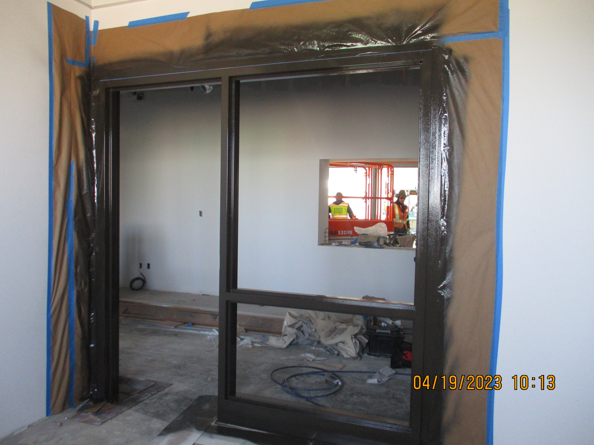 An interior construction site with a plastic-covered doorframe, tools on the floor, and a reflection of two workers.