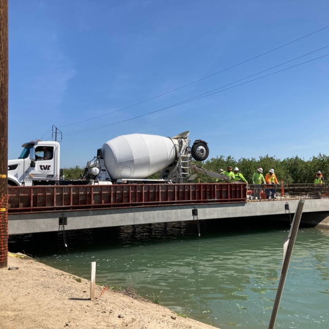 Construction workers, cement mixer truck, and partially constructed bridge over water.