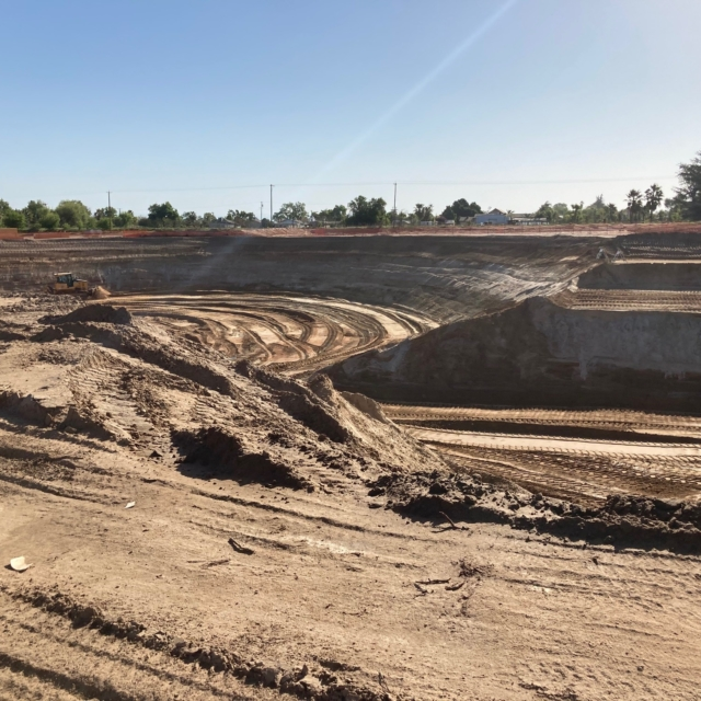 Large construction excavation site with tire tracks, heavy machinery in the distance under a clear sky.