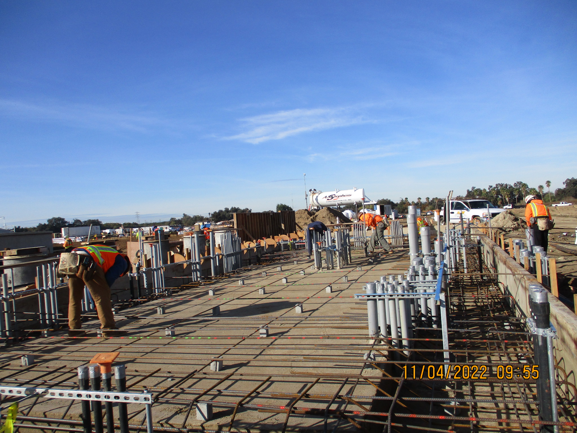 Construction site with workers, rebar framework, concrete forms, and a mixer truck under a clear sky.