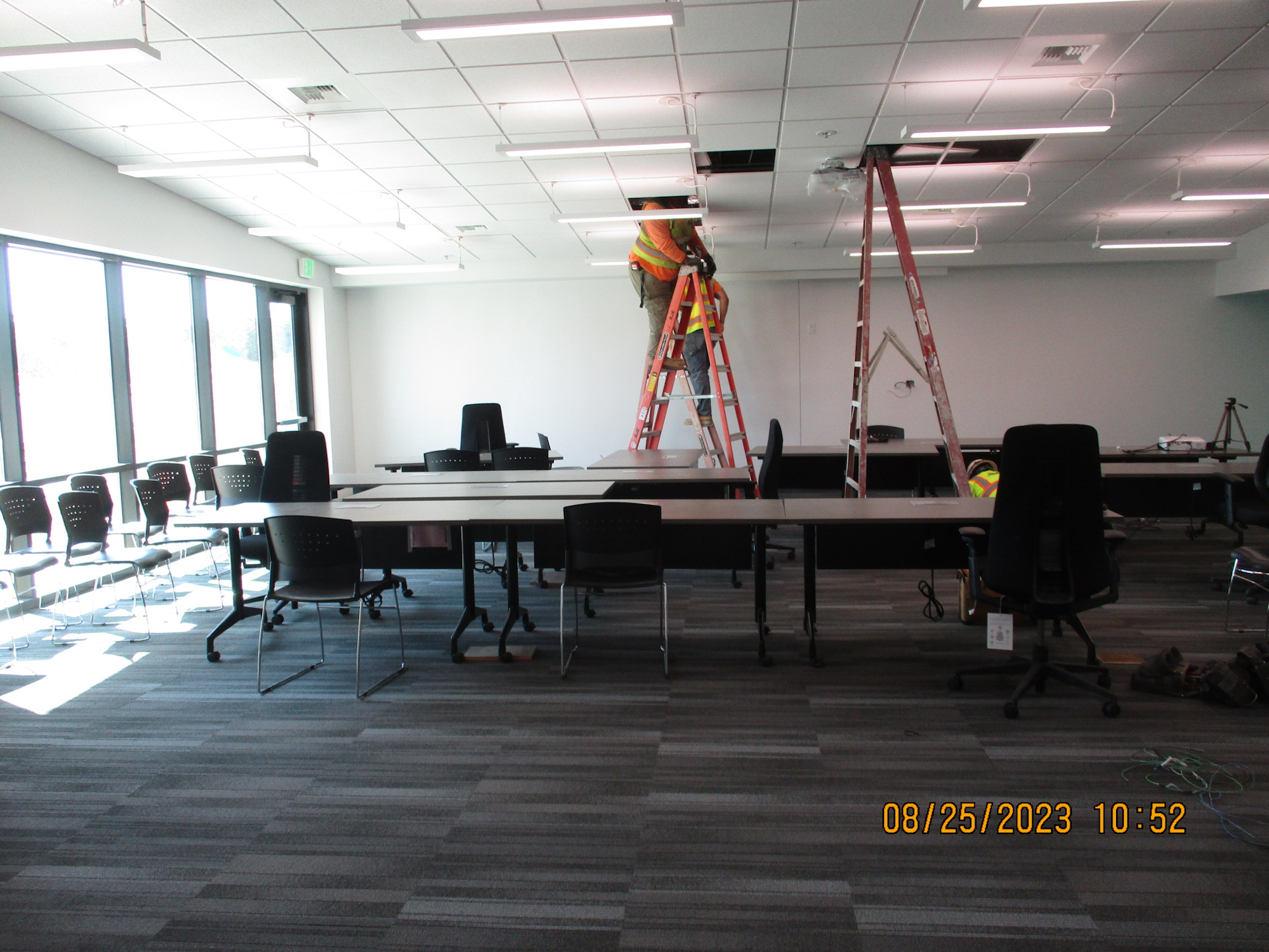 Conference room with tables and chairs, two workers on ladders installing fixtures.