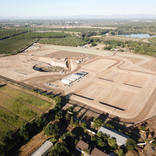 Aerial view of a construction site with cleared land and dirt roads, surrounded by greenery and a water body nearby.