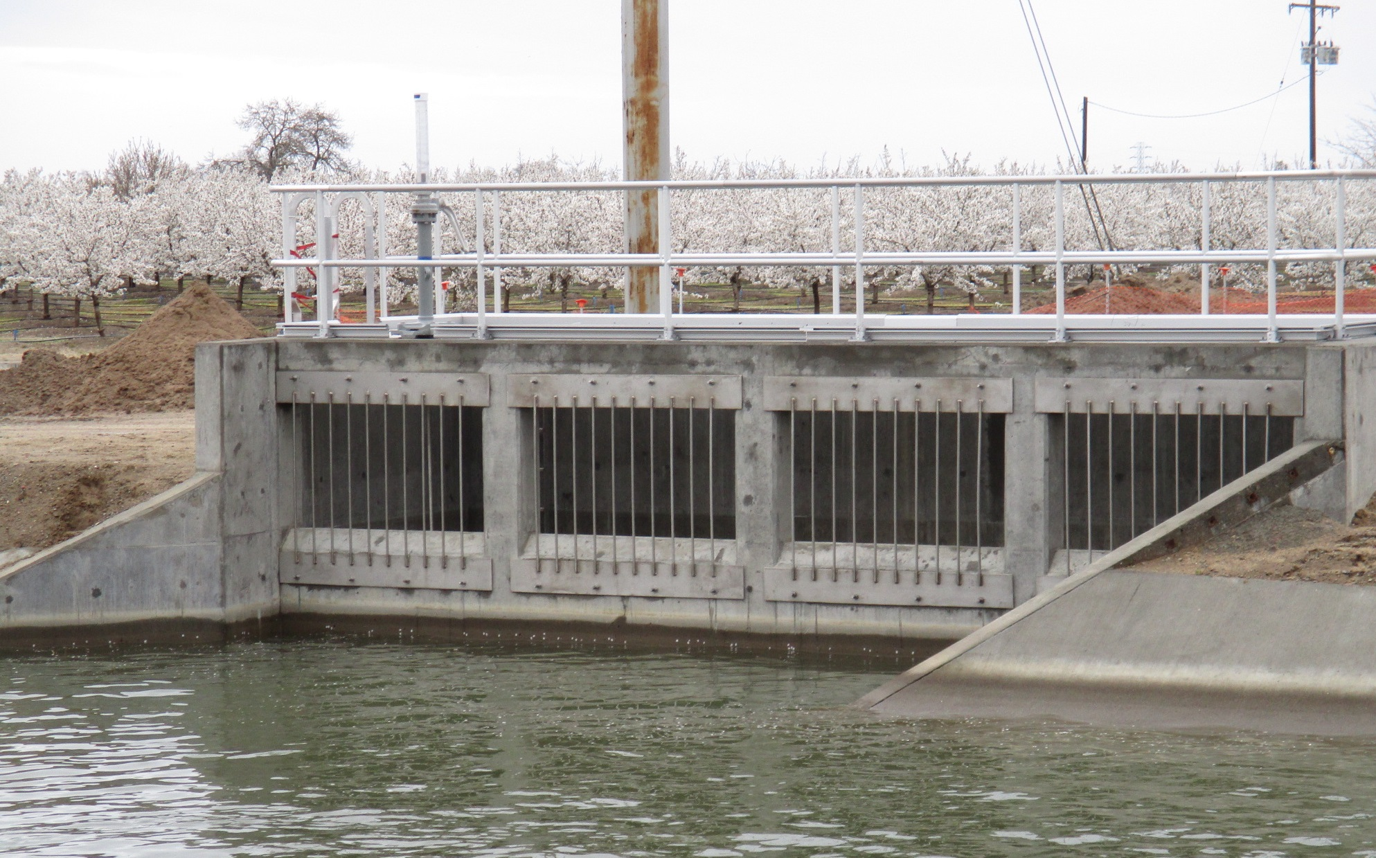 Water gate with metal grates, concrete structure, flowering trees in the background.