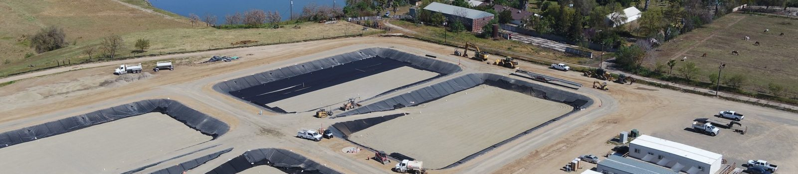 Aerial view of a construction site with two large excavated areas, likely foundations, heavy machinery, and some vehicles.