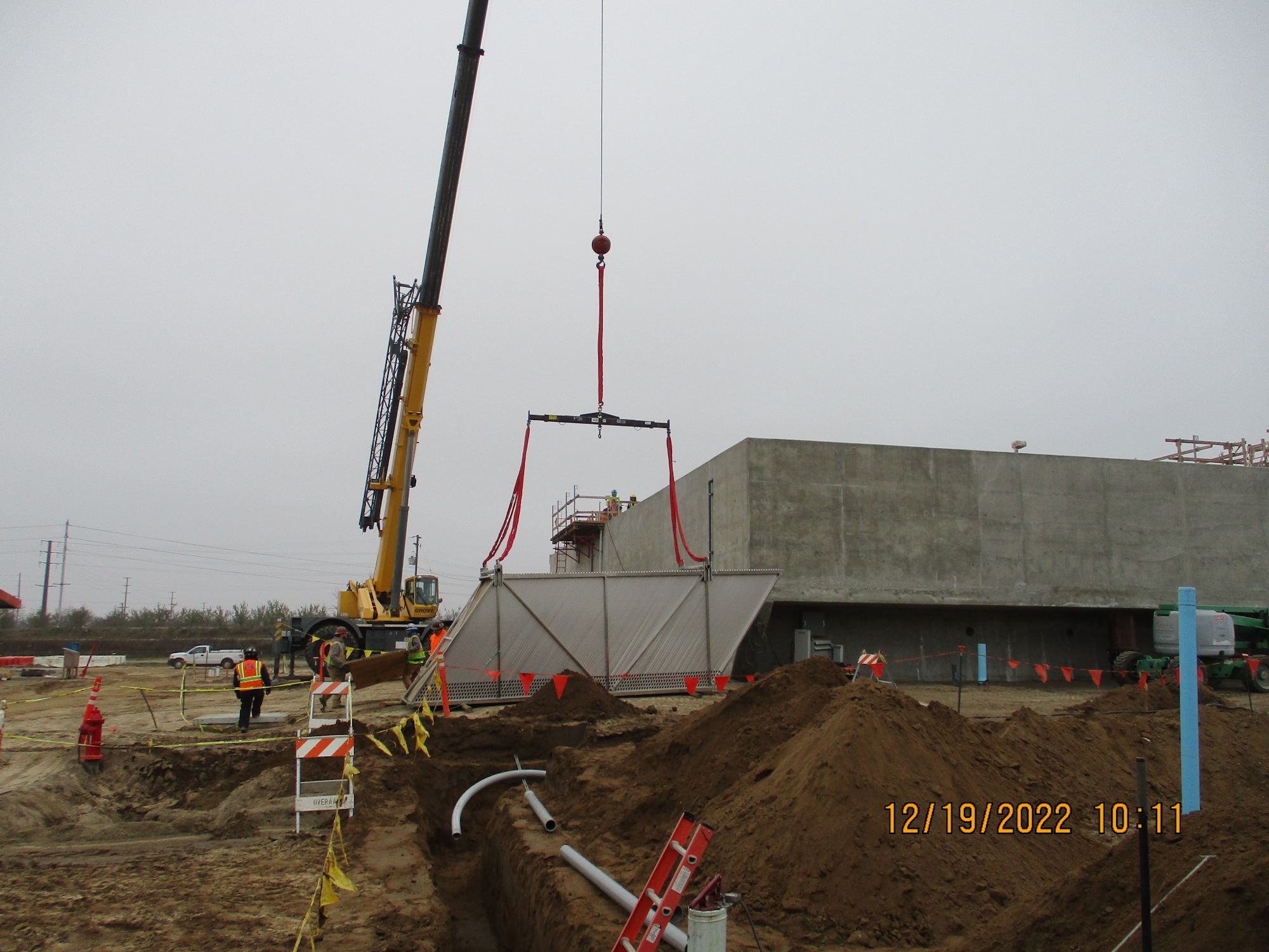 A crane lifting a large object at a construction site with workers and safety barriers visible.